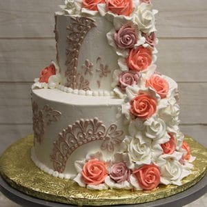 The WC-000E is a two-tiered white cake with lace-like piping and cascading buttercream roses in pink, coral, and white, presented on a gold-covered cake board atop a gray cake stand.