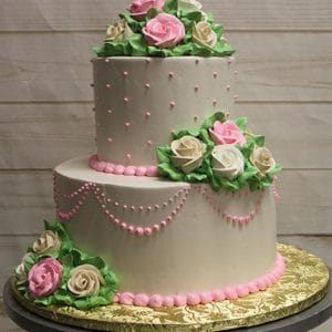 The WC-000H is a two-tiered white cake adorned with pink and white icing roses, green leaves, and pink decorative dots and swags. It is displayed on a round gold board against a light wood background.