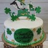 The BD-116 is a round cake with green shamrocks, gold dots, and a leprechaun mascot topper. A green plaque says "Happy 60th Birthday Papdi." The cake sits on a gold tray trimmed with green beads.
