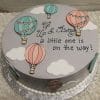 BB-065 is a round cake with pastel hot air balloons, white clouds, and the message “Up & Away a little one is on the way!” on light gray fondant—perfect for baby showers.