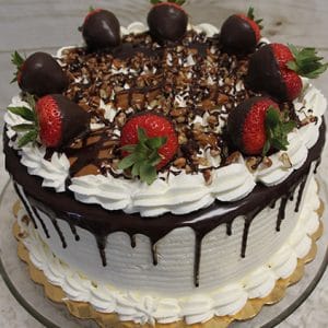 The BD-015 Chocolate Turtle Cake is a round cake with white frosting, chocolate ganache drips, whipped cream borders, and is topped with chocolate-covered strawberries, nuts, and chocolate drizzle on a glass stand.
