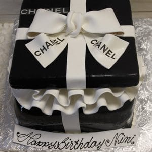 The BD-027E Gift Box Black is a square black cake styled as a Chanel gift box, featuring a white fondant bow, CHANEL on the ribbons, and a "Happy Birthday Nimi" plaque at the base.