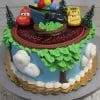 The SC-010 Cars cake is round with toy cars, trees, and balloons on top, green and blue landscape icing, "Happy Birthday" in red icing, and clouds, trees, and grass decorating the sides.
