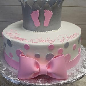 The BB-028 cake is round with pink and gray polka dots, a pink bow, a silver crown topper with pink baby footprints, and pink script text on the top edge. It sits on a silver board.