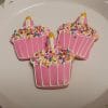 The CE-001b Birthday features three pink cupcake-shaped cookies topped with white icing, colorful sprinkles, and a small yellow candle, all arranged on a white decorative plate.