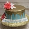 The BD-057B Gold Drip Aqua is a round cake with blue icing, gold drip topping, and large pink and white flowers, displayed on a gold foil board. A banner reads “Happy Birthday Mike & Melina” in blue script.