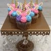 Six CC-021 Chocolate with white filling cupcakes, topped with pink, blue, and purple swirled frosting plus gold horns and fondant ears, are displayed on an ornate brown cake stand adorned with crystal-like beads.