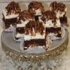 Six Turtle Squares topped with white frosting and caramel drizzle are displayed on an ornate silver cake stand with decorative chains, set against a textured white background.