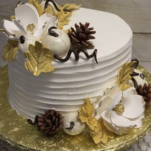 BD-130 is a white frosted cake adorned with white flowers, gold leaves, pinecones, and small white pumpkins on a gold cake board against a textured background.