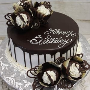 BD-057A Ganache Drip: A round chocolate birthday cake with white icing, chocolate drip, "Happy Birthday" on top, and adorned with decorative chocolate and cream flowers. The cake board features a floral-patterned foil wrap.