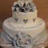 TC-139: A two-tier white and silver cake with ruffled accents, heart motifs, elegant piping, a fondant flower, and a large silver “25” topper—perfect for celebrating a 25th anniversary.