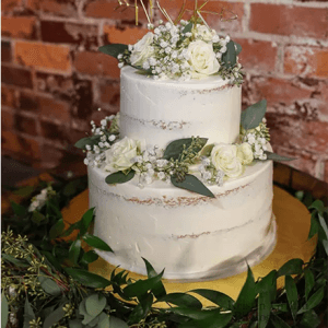 The WD-000A is a two-tiered white wedding cake with semi-naked frosting, adorned with white roses, greenery, and baby's breath, finished with a gold N♥K topper and surrounded by lush leafy greenery against a brick wall.