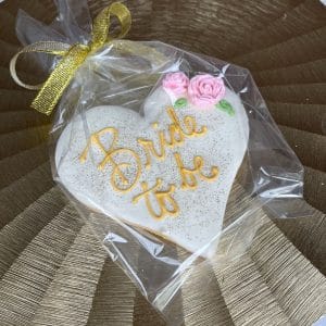 The CE-003g bridal is a heart-shaped cookie with "Bride to be" in yellow icing, pink floral decorations, wrapped in clear plastic with a gold ribbon, and placed on a textured gold placemat.