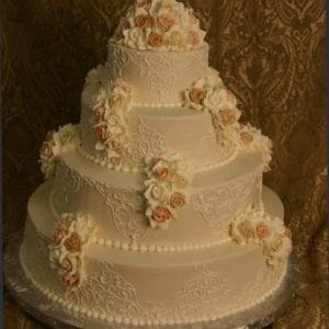 The WC-100 is a four-tiered wedding cake with intricate white icing and cream and pale pink sugar roses, displayed against a brown patterned background.