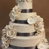 WD-025 is a four-tier ivory wedding cake adorned with large ivory sugar flowers, dark satin ribbon bands, and intricate piped swirl designs, beautifully set against a brown background.