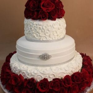 WD-036 is a two-tier white wedding cake with intricate swirls, silver accents, and a bouquet of red roses on top. Red roses surround the base, all beautifully set against a brown background.