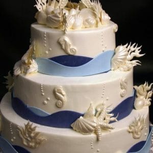 The WD-053 is a three-tiered white cake adorned with sea-themed fondant, featuring seahorses, seashells, coral accents, and blue wave bands around each tier.