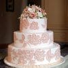 The WD-102 is a three-tiered white cake featuring blush pink floral lace designs and a bouquet of pink and ivory sugar flowers on top, elegantly displayed on a round platter.