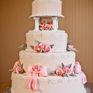 The WD-138 is a four-tiered white wedding cake adorned with pink fondant bows, roses, green leaves, and ornate white icing details, presented on a silver cake board.