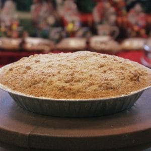 A Pie-Blueberry with a golden brown crumb topping rests in a foil pie tin on a round surface, with blurred festive decorations in the background.