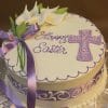 EA-015 is a round Easter cake with ornate white and purple icing, decorated with a purple ribbon, white calla lilies, a purple cross, and "Happy Easter" written in purple icing.