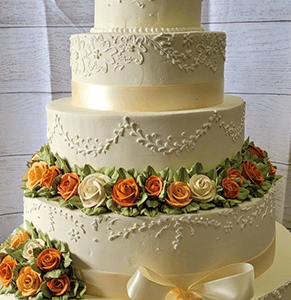 WC-052 is a four-tiered white cake adorned with intricate icing, orange and white rose clusters, green leaves, and a large cream-colored ribbon bow on the bottom tier.