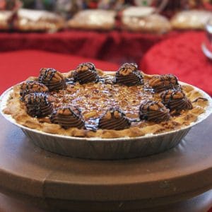 Pie-Pecan sits on a round wooden surface, topped with chocolate cream swirls, drizzled with chocolate sauce, and sprinkled with nuts. The background is blurred with red and festive decorations.