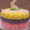 The EA-018 is a round cake with yellow, purple, and pink rosettes, topped with a white chocolate bunny and a “Happy Easter” sign in pink icing. It sits on a foil-covered board.