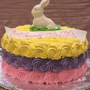 The EA-018 is a round cake with yellow, purple, and pink rosettes, topped with a white chocolate bunny and a “Happy Easter” sign in pink icing. It sits on a foil-covered board.