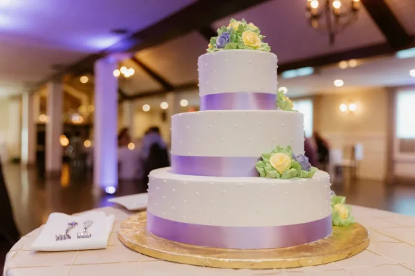 A three-tiered white Konditor Meister cake decorated with small dots, wrapped in light purple ribbon, and topped with yellow and green flowers, displayed on a golden base in a warmly lit room.