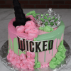 CB-040 is a round cake with pink and green frosting, topped with a black witch hat, silver tiara, and a “WICKED” sign on the side, inspired by the musical Wicked. Presented on a silver board.